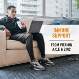 Hombre sentado en sofá bebiendo proteína en polvo, usando laptop, texto de soporte inmune.